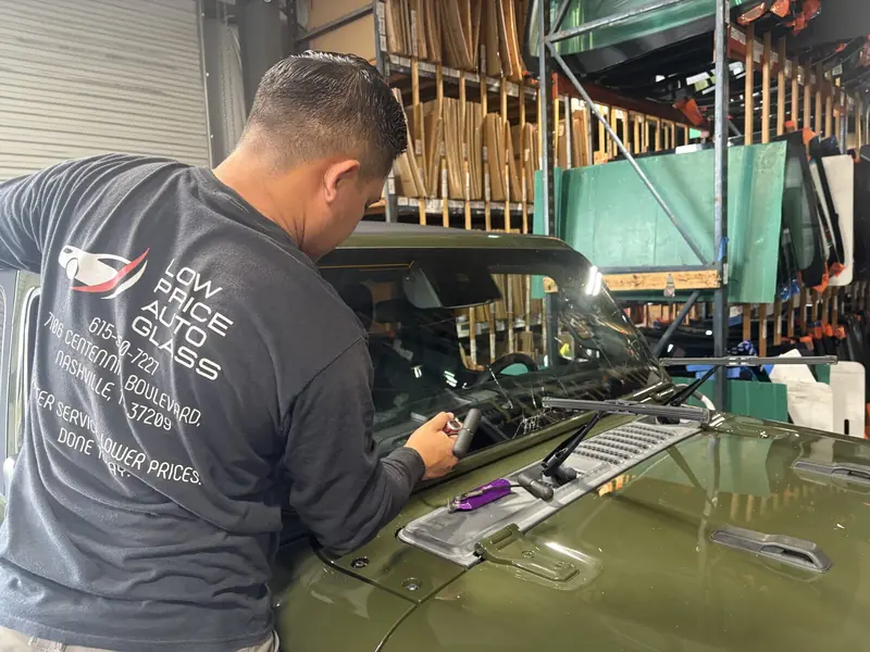 A Low Price Auto Glass technician in a company shirt installing a windshield on an olive green Jeep